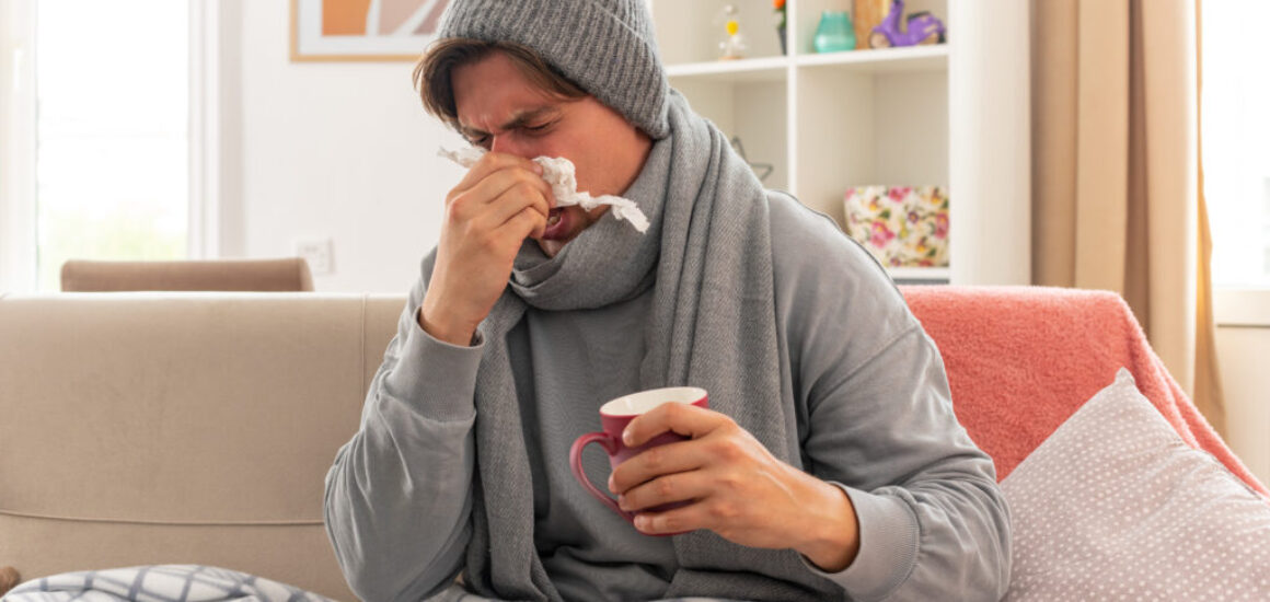 unpleased young ill slavic man with scarf around neck wearing winter hat wipes his nose with tissue and holds cup sitting on couch at living room