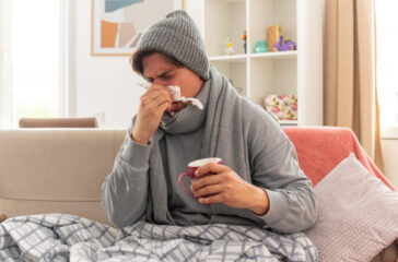 unpleased young ill slavic man with scarf around neck wearing winter hat wipes his nose with tissue and holds cup sitting on couch at living room