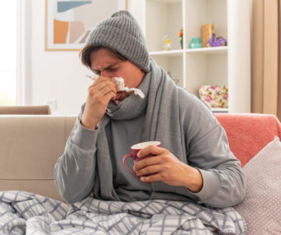 unpleased young ill slavic man with scarf around neck wearing winter hat wipes his nose with tissue and holds cup sitting on couch at living room